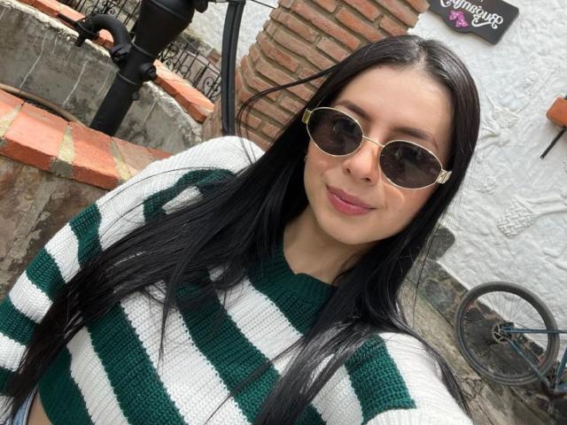 IsabellaJames with long black hair, wearing green and white striped sweater, brown aviator sunglasses, smiling, standing outside with brick wall and black pole in background.