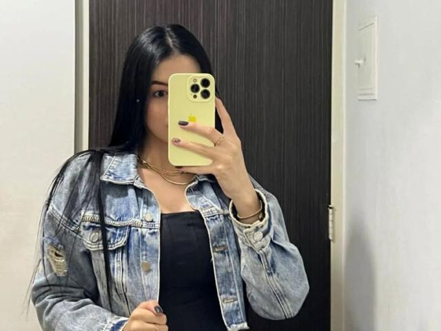 IsabellaJames with long black hair, wearing a denim jacket over a black top, holding a yellow iPhone, taking a mirror selfie, showing off her nails and necklace.
