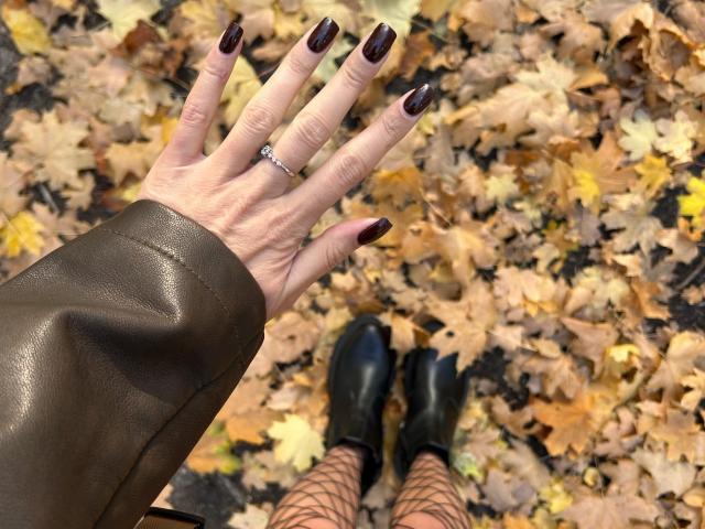 Karish's hand with dark purple nails, a silver ring, and a brown leather sleeve, standing on autumn leaves, wearing black boots and fishnet stockings, photo.
