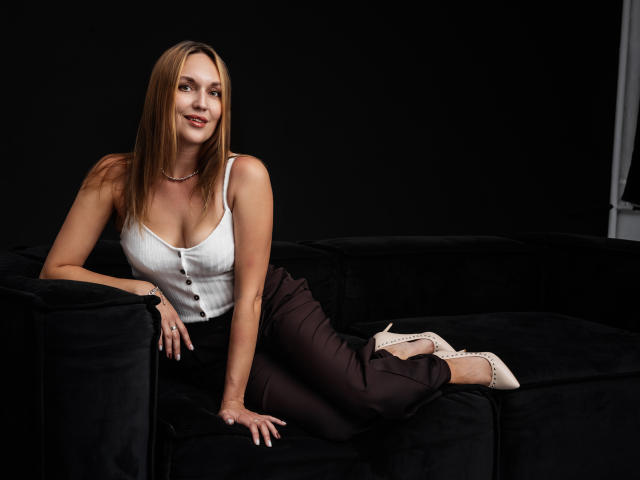 EvelynCute with long brown hair, white tank top showing small tits, brown pants, and pink heels, lounging on a black couch, smiling, hand on seat.