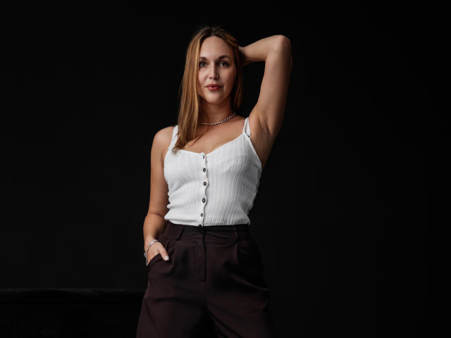 EvelynCute with brown hair, white ribbed tank top, buttoned down, and brown pants, hand in pocket, other arm raised, dark background, smiling, small tits, cute.