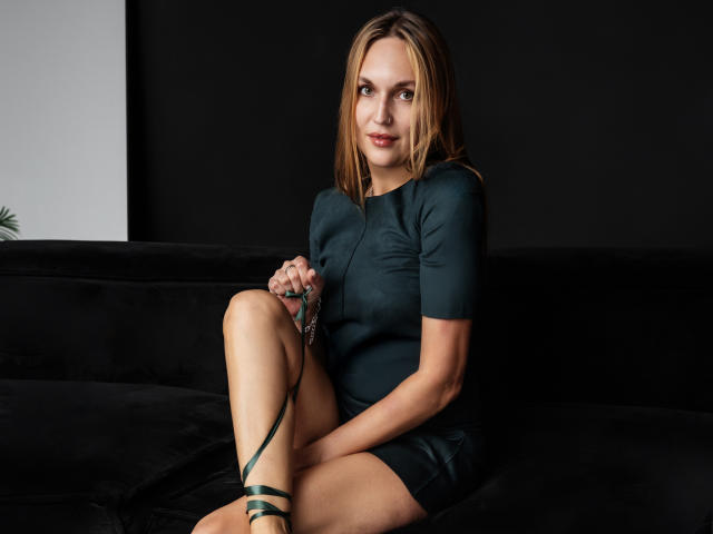 EvelynCute with blonde hair, green dress, and green strappy heels, sitting on a black couch, holding a green leash, smiling at the camera, showing her cute ass.