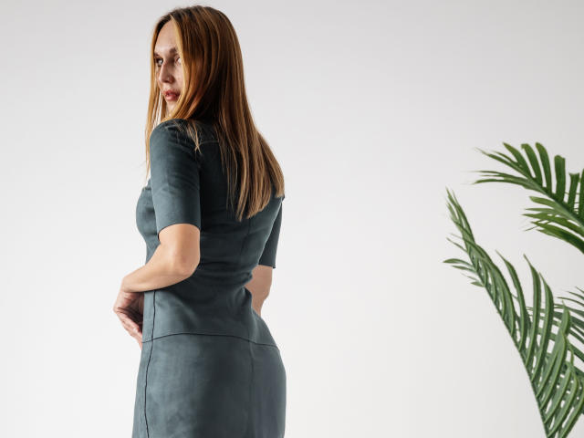 EvelynCute, with long brown hair, wearing a tight grey dress, showing her ass, standing with hands on her hips, looking over her shoulder, next to a green plant.