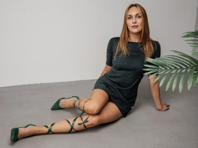 EvelynCute sits on a gray floor, wearing a dark green, short dress and matching green shoes with lace-up details. Her straight, light brown hair falls to her shoulders. She leans on her right arm, with her left arm resting on her knee. A green potted plant is on the right. Her expression is neutral, and she looks slightly to the left. The image has a clean, minimalistic style with a focus on her green outfit and relaxed pose.