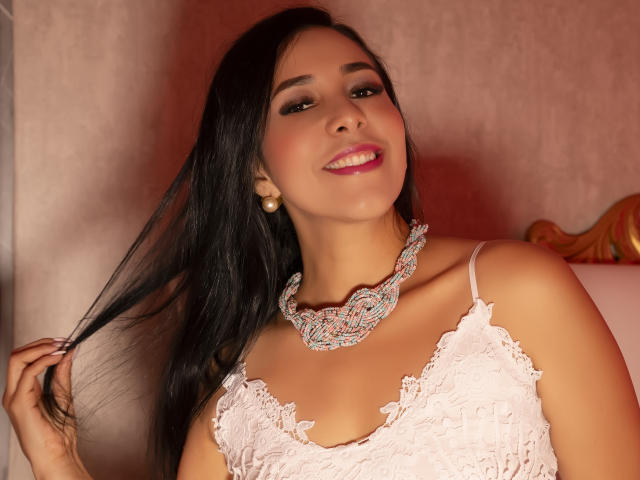 Katty Rivera, con lunghi capelli neri, sorridente, indossa una blusa di pizzo rosa e una collana vistosa con gioielli, si tiene i capelli, mostra il seno e indossa orecchini di perle..