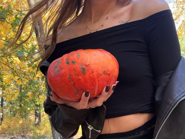 Karish holding a large, orange, slightly spotted pumpkin with her dark-nailed hand. She's wearing a black off-shoulder top and a black jacket, with autumn leaves in the background.