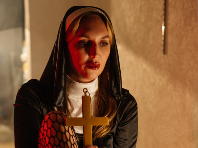 EvelynCute, dressed in a black nun's habit with a white wimple, holds a golden cross. Red light casts a shadow on her face, contrasting with her blonde hair and red lipstick. She wears fishnet gloves on her right arm. The textured wall in the background is illuminated by a single light source. The composition is intense and dramatic.