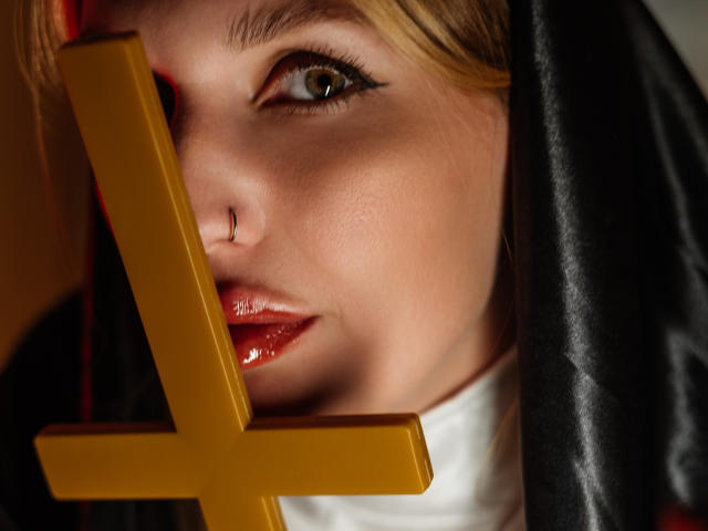 Close-up of EvelynCute with blonde hair, green eyes, and glossy red lips. She holds a yellow cross in front of her face, partially obscuring it. Her skin is smooth, and she wears a black veil. The cross's yellow color contrasts with the black veil and white collar. The cross is in the foreground, centered. The lighting highlights her facial features and shiny lips. The overall composition is intense and focused on EvelynCute's face.