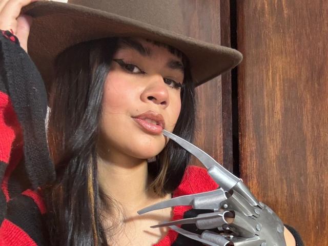 IsabellaMarynn wears a brown hat and a red and black striped sweater. She has long black hair with bangs and dark eyebrows. She holds a metallic, claw-like hand near her lips. Her lips are slightly parted, showing a hint of pink lipstick. The texture of the hat is rough, and the wooden background is visible. The image has a realistic, photographic style. Her expression is neutral. The claw hand is detailed, with sharp, metal-like fingers.