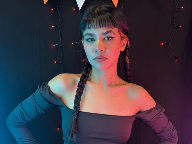 IsabellaMarynn ha i capelli castano intrecciati, una maglietta nera con le spalle scoperte e eyeliner rosso, ed è in piedi con le mani sui fianchi, con delle luci decorative rosse che brillano sullo sfondo..