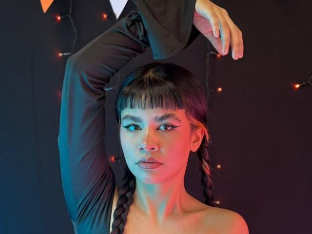 IsabellaMarynn, con i capelli scuri intrecciati, indossa una maglia nera a maniche lunghe, con un braccio alzato, mostrando il viso e la parte superiore del corpo, illuminata da luci blu e rosse, con un'espressione seria..