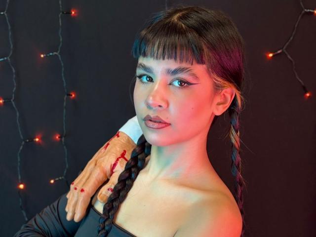 IsabellaMarynn with braided hair, wearing off-shoulder black top, has bloody hand on her neck, red lips, dark makeup, and string lights in background, looking serious.