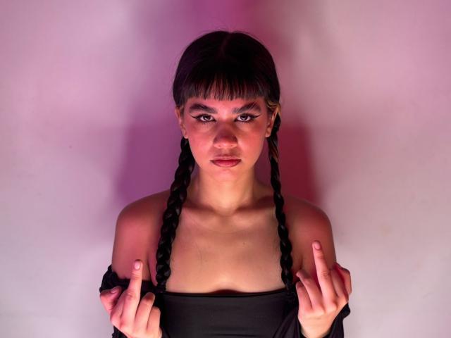 IsabellaMarynn with braided black hair, dark off-shoulder top, and pinkish-purple lighting, holding up two fingers, showing off her tits, with bold winged eyeliner and pink lipstick.