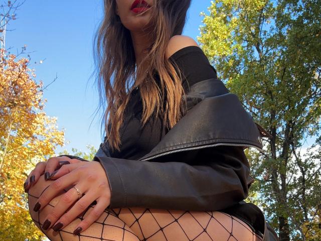Karish with long brown hair, red lipstick, black off-shoulder top, black leather jacket, fishnet stockings, and dark nail polish, squatting outdoors, showing her thighs and ring.