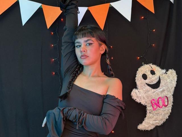 IsabellaMarynn, con i capelli scuri intrecciati, un abito nero a spalle scoperte e guanti neri, alza un braccio, in piedi accanto a un fantasma di peluche bianco inquietante con la scritta "BOO" in rosa..