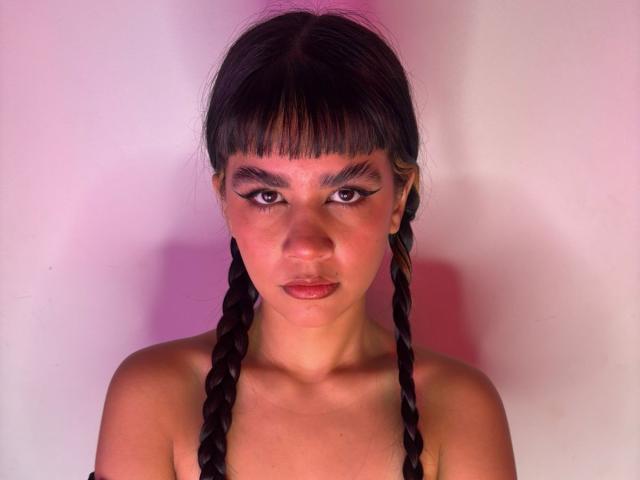 IsabellaMarynn has a serious expression with dark, full eyebrows and large, black eyeliner accentuating her eyes. She has straight, black hair in two braids, with bangs covering her forehead. Her skin is medium brown and smooth. She is topless, with her shoulders visible. The pink and purple lighting creates a soft, warm glow on her face. The image is centered with IsabellaMarynn's face filling the frame. The composition is straightforward, focusing on her face and braids.