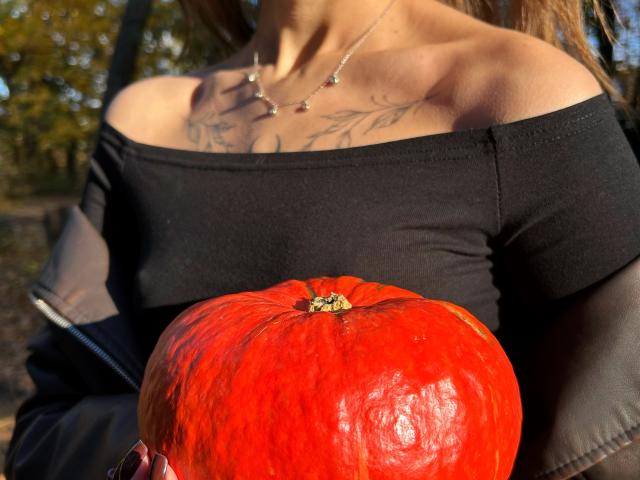 Karish with tits visible, black off-shoulder top, holding a red pumpkin, tattoos on chest, necklace, sunlit, brown hair, black jacket, outdoor setting.