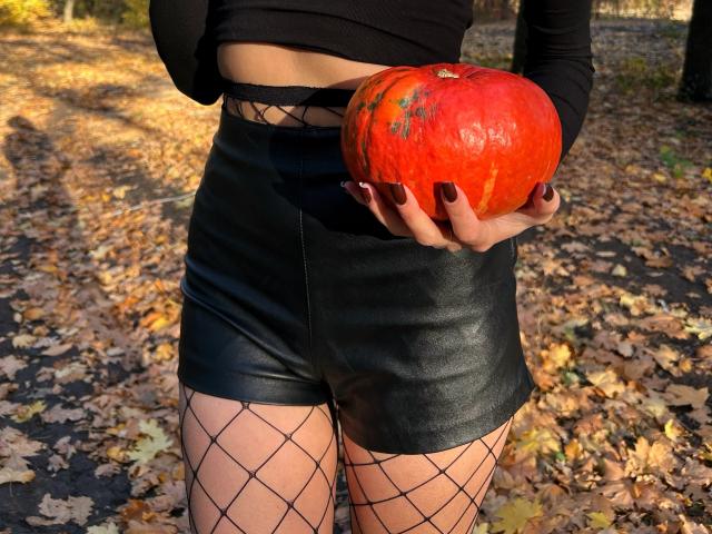 Karish holding a big orange pumpkin in black leather shorts and fishnet stockings, her dark nails contrasting with the bright pumpkin. Autumn leaves on the ground.