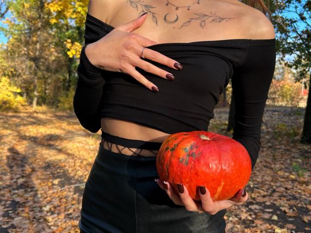 Karish sosteniendo una calabaza roja con la mano sobre sus senos, vistiendo una blusa negra con un hombro descubierto y una falda negra, en un bosque otoñal iluminado por el sol..