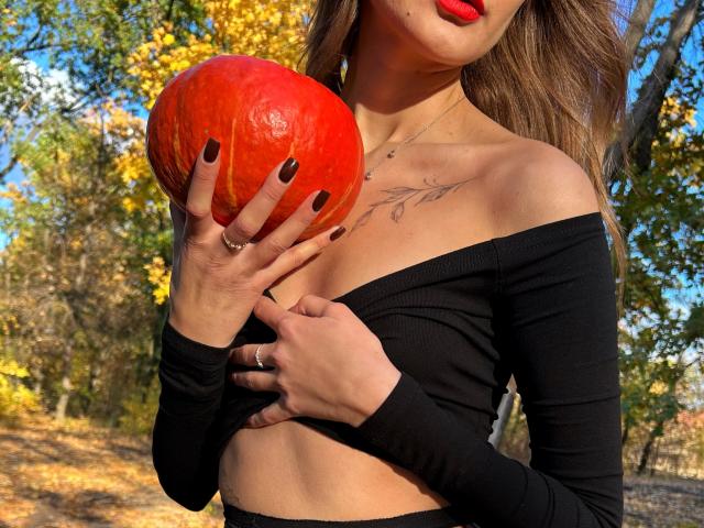 Karish holding a big red pumpkin, wearing a black off-shoulder top, red lipstick, black nails, and a tattoo above her tits, with her hand on her pussy.