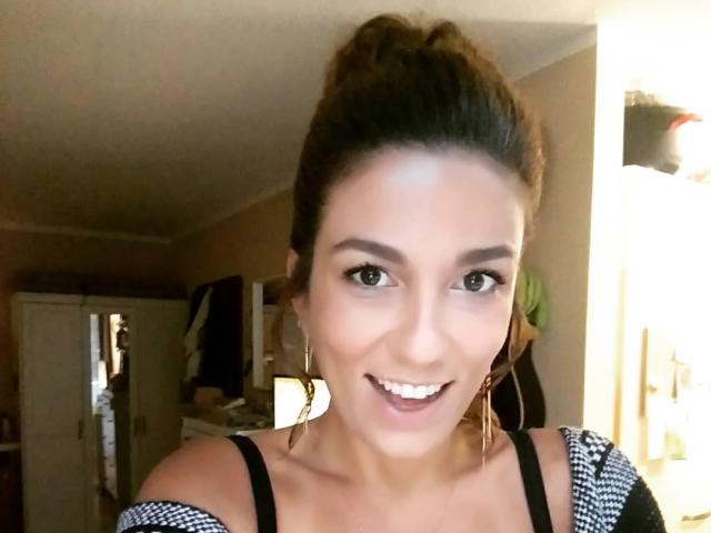 OliviaLux tiene la piel morena, ojos marrones, y está sonriendo con la boca abierta, llevando aretes grandes y una blusa blanco y negro, con su cabello castaño recogido en una coleta alta..
