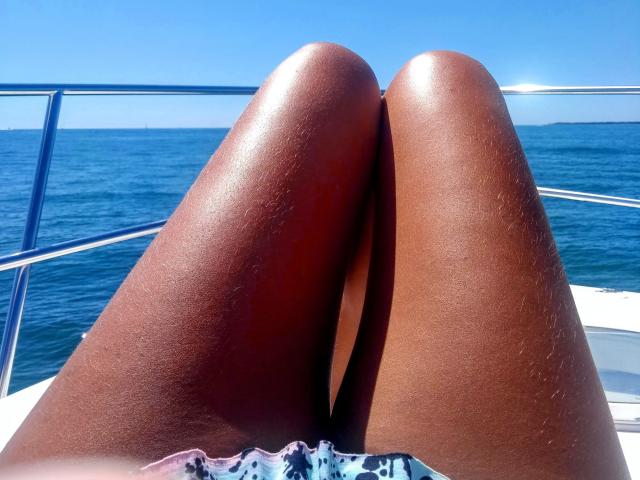 Le cosce marroni e lucide di OliviaLux sono divaricate su una barca, con l'oceano blu e il cielo sereno sullo sfondo. Le sue ginocchia sono più vicine alla macchina fotografica..