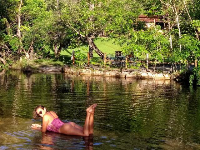 OliviaLux è sdraiata in uno stagno poco profondo, indossando un bikini rosa in pizzo, con i glutei sollevati e le gambe piegate, e guarda indietro con un sorriso. I suoi capelli castani sono bagnati..