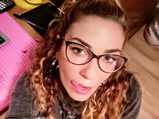OliviaLux, con i capelli castano ricci, gli occhiali e il rossetto rosa, con la lingua leggermente fuori, che guarda verso l'alto la fotocamera, indossa una maglietta grigia, su un pavimento di legno..