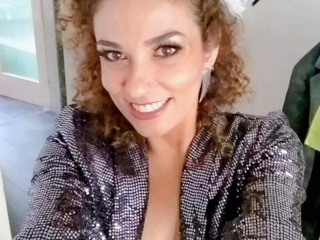 OliviaLux with curly brown hair, smiling, wearing a sparkly black and silver jacket, showing a hint of her boobs, wearing diamond earrings, in a selfie.