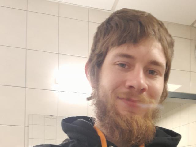 Ozzachy has a brown beard and mustache, wavy brown hair, and a slight smile, wearing a black hoodie with orange trim, in a blurry selfie.