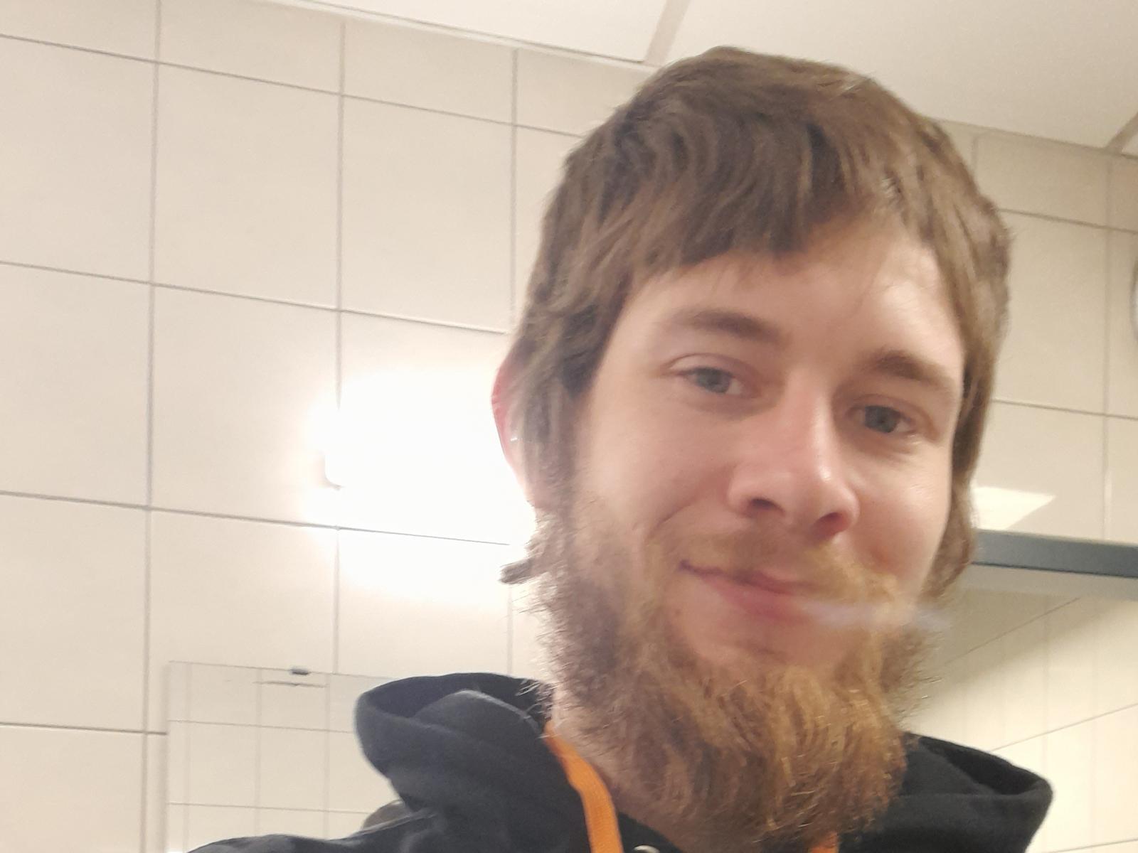 Ozzachy hat einen braunen Bart und Schnurrbart, welliges braunes Haar und ein leichtes Lächeln. Er trägt eine schwarze Kapuzenjacke mit orangefarbenen Akzenten und ist auf einem unscharfen Selfie zu sehen..
