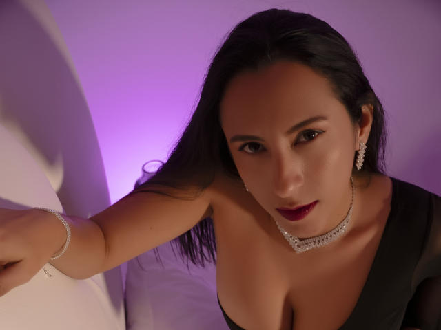 JadeFoster with dark hair, wearing a black dress, choker, and earrings, looking up with red lipstick, her tits visible, purple lighting, and a bracelet on her right arm.