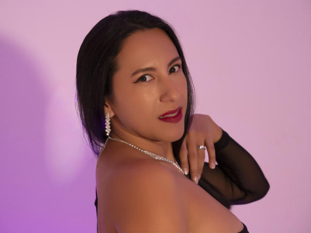 JadeFoster with dark hair, red lipstick, pearl earrings, and a black sheer sleeve, looking over her shoulder, showing her tits and a ring on her hand.