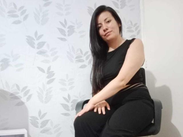 MargarettCooper with long black hair, black sleeveless top, and black pants, sitting on a chair, hands on knees, looking slightly to the side, in a simple room.
