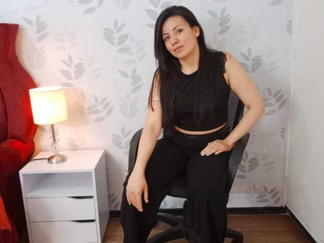MargarettCooper with long black hair, wearing a black sleeveless crop top and black pants, sitting on a gray chair, hand on knee, red chair and white lamp in background.