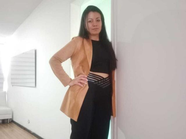 MargarettCooper with long black hair, tan jacket, black top, and black pants with strappy waist, standing against white wall, hand on hip, looking straight at camera.