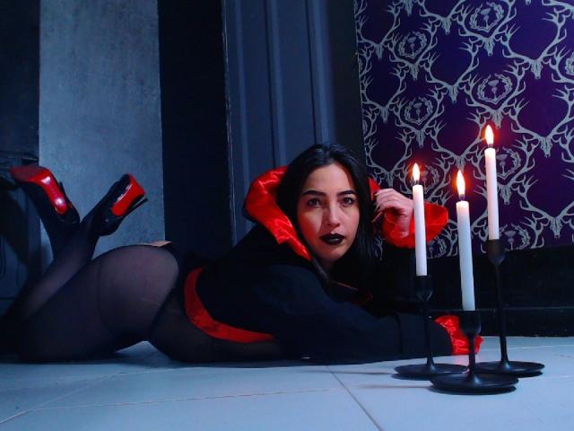 RoseChapman lying on white tile, black sheer pantyhose, red high heels, red satin hood, black lipstick, black eyes, red belt, black top, candles with flames in front.