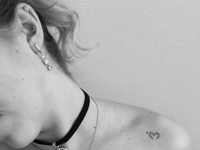 SugarrX's ear with a silver stud and a small heart tattoo on her shoulder, wearing a black strap and a thin necklace, in a black-and-white photo.