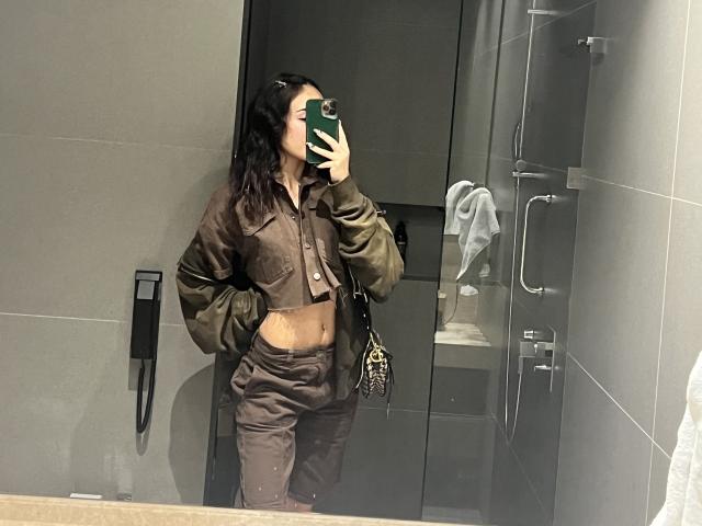 AitanaJohns with dark hair, wearing a brown jacket and matching pants, showing her midriff, holding a green phone, standing in a gray-tiled bathroom, reflection in mirror.