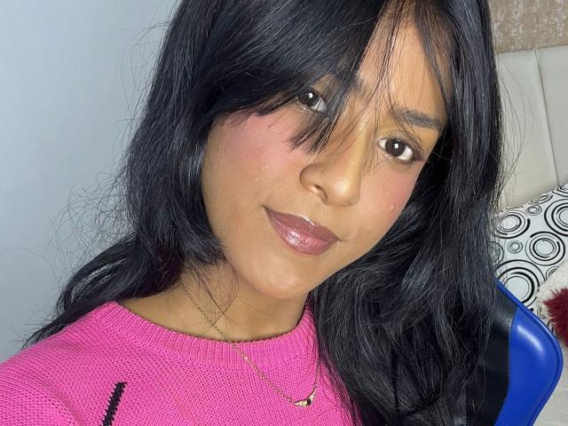 Babilett with black hair, pink sweater, and shiny pink lipstick, smiling at the camera with a slight blush. Her brown eyes and subtle makeup are visible.
