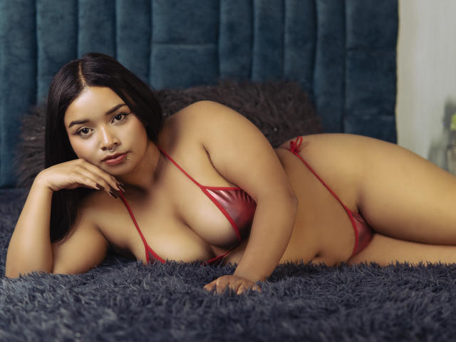MiaDuvall is lying on a bed in red, shiny, barely-there bikini, showing her big tits and ass, with a relaxed, sultry expression, on a fluffy gray blanket.