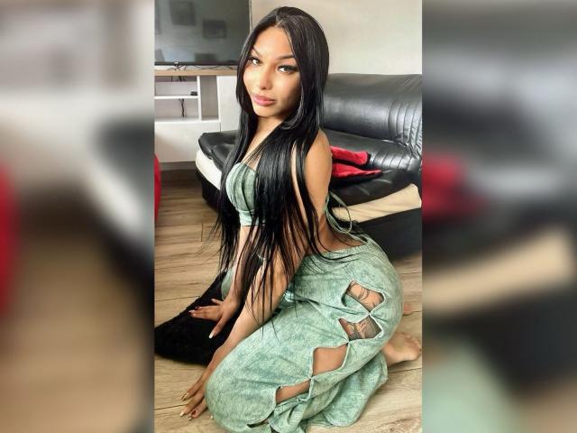 Amaranta69s with long black hair, green eyes, and tan skin, kneeling in a green, torn, sleeveless top and pants, showing her ass and small, round boobs.