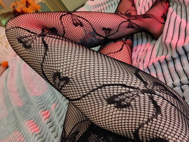 BerryDiva's legs in black fishnet stockings with floral patterns, showing her sexy thighs and red-lit skin, lying on a striped pink and gray blanket.