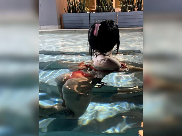 Amaranta69s with wet black hair, pink hairclip, red bikini, and visible ass, kneeling in a pool, back to camera, water reflecting sunlight.