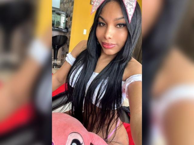 Amaranta69s with long black hair, pink bow, and white off-shoulder top, showing her tits, sitting on a red chair, smiling at the camera. Her pussy is barely visible.