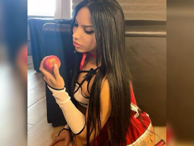 Amaranta69s with long black hair, holding a red apple, wearing a red and black strappy top, white arm sleeve, kneeling, looking down, in a room.