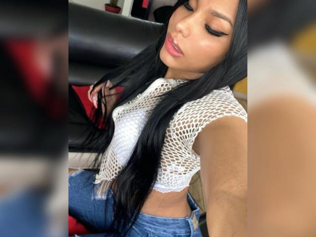 Amaranta69s with long black hair, wearing a white fishnet top and blue jeans, has her eyes closed, pink lips slightly parted, showing her tits and a cute face.