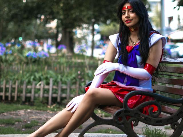 VioletXBloom sits on a wooden bench in a park, dressed as a Sailor Moon. She has long black hair, red lipstick, and wears a red headband with a blue and white dress, red skirt, and white gloves. Her crossed legs show smooth, tan skin. She has red arm cuffs and a red heart-shaped brooch. The bench has an ornate black metal frame. Her expression is calm and slightly confident. The image has a natural, outdoor setting with blurred greenery and flowers in the background.