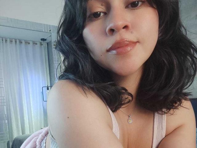 JuliettaHott has shiny, glossy lips and wavy black hair. She's wearing a pink tank top with a daisy necklace. Her tits are partially visible. Her skin is light.