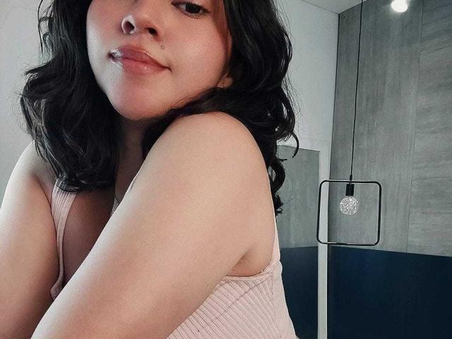 JuliettaHott with wavy black hair, pink ribbed tank top, showing her tits, smiling slightly, light skin, in a modern room with a hanging light bulb.